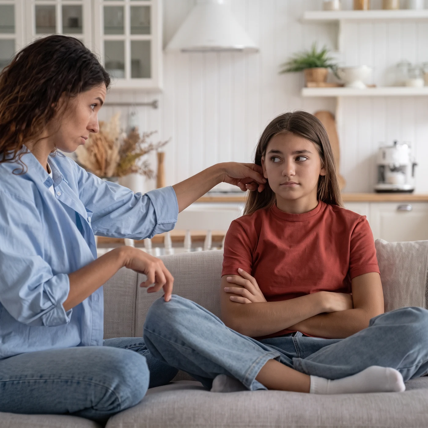 A mother on the sofa reaches toward her teenage daughter, who sits with her arms crossed, looking away
