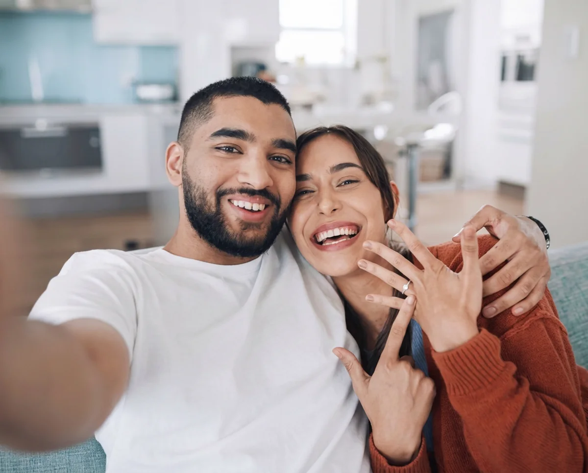 A just-engaged couple taking a selfie on their sofa at home, laughing, she holds up her hand to show the ring